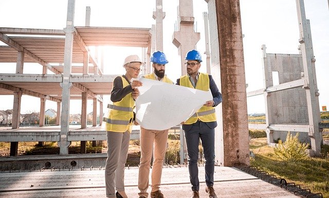Three construction workers in yellow vests and hardhats stand in front of a partially built development and review a building plan]